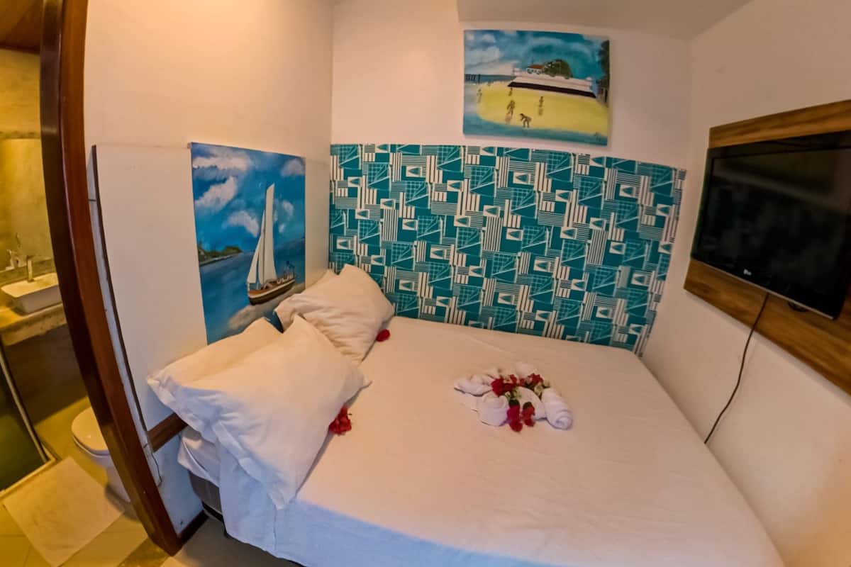 Ett hotellrum med dusch, en säng med ett vitt överkast, en målning av en segelbåt och en sänggavel med mönster.