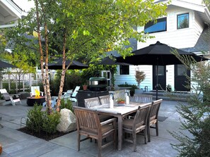 A patio with a dining table, chairs, and a grill. There are umbrellas providing shade, and a fire pit is visible. - Sparkling Clean, Chemical Free Ultra-Modern Bay View Loft. Superb Location.  (Tacoma)