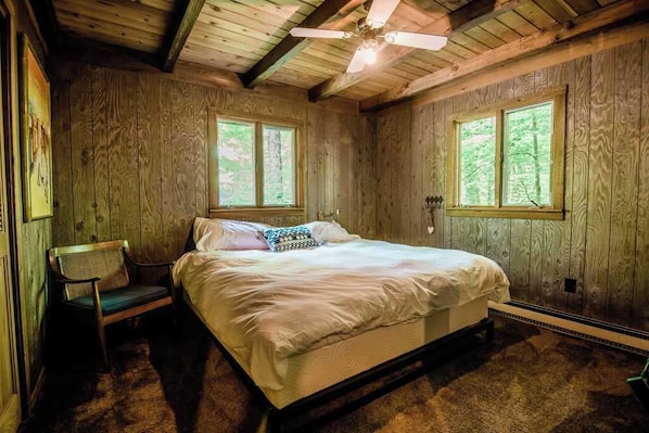 A wooden cabin bedroom with a bed, two windows, a ceiling fan, and a chair.