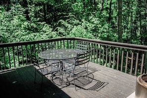 A wooden deck with a metal table and chairs, surrounded by dense green foliage.