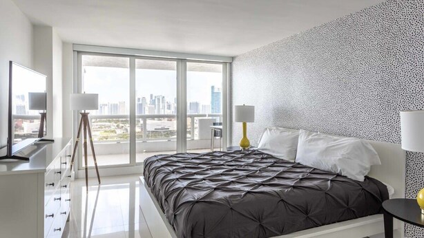 A modern bedroom with a large bed, a city view, and a white dresser.