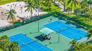 An outdoor tennis court with a green roof, surrounded by palm trees and a sandy area.