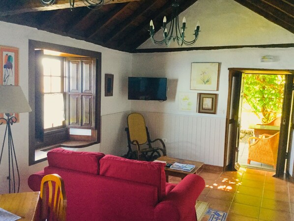 Una sala de estar acogedora con un sofá rojo, una silla de madera, un televisor montado en la pared y una puerta que da a un jardín.