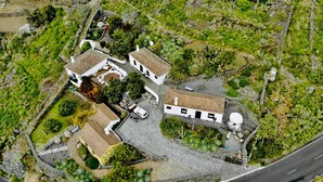 An aerial view of a property with multiple buildings, including a large white structure with a tiled roof and a smaller building with a similar roof. There are paved areas and greenery surrounding the buildings.