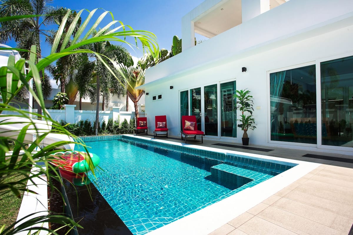 Un espace piscine moderne avec une piscine à l’eau d’un bleu limpide, des chaises rouges et un bâtiment blanc doté de grandes fenêtres.