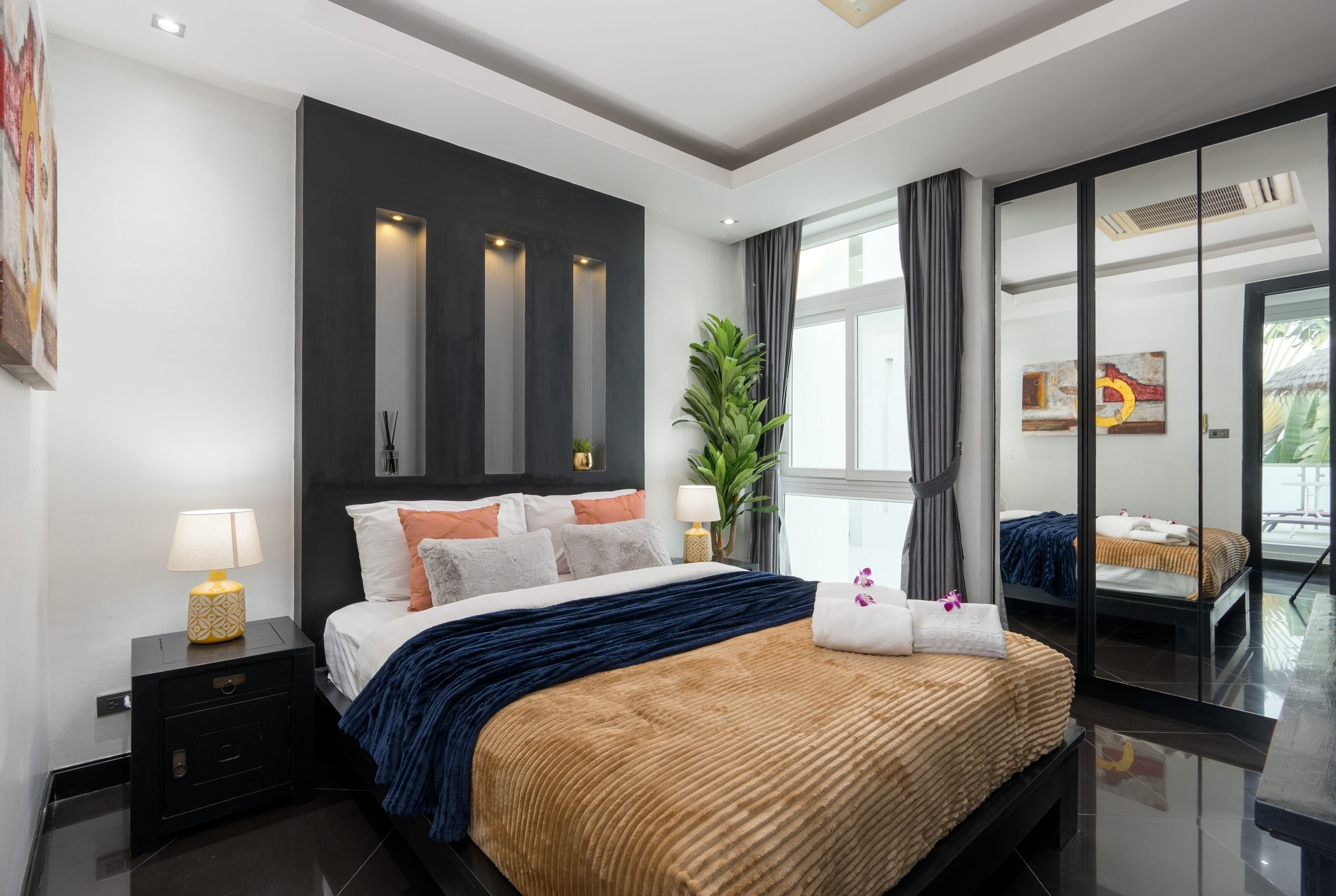 Een moderne hotelkamer met een groot bed, een nachtkastje met lamp, een potplant en een grote spiegel.