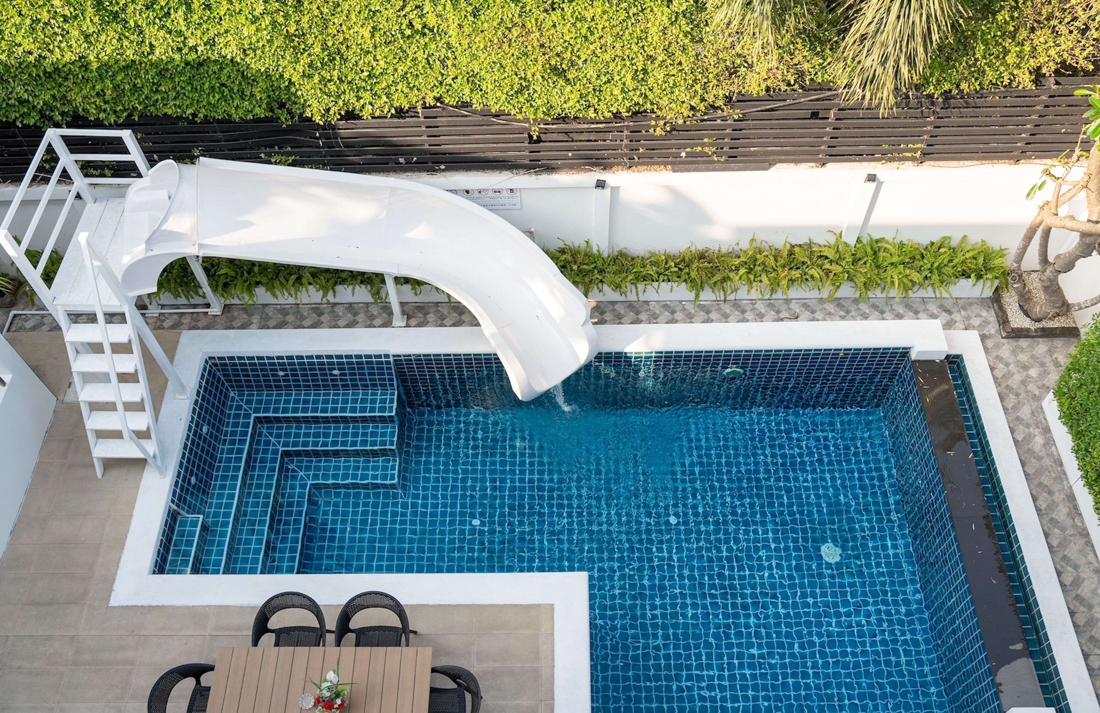 A swimming pool with a white slide, surrounded by a white wall and a wooden deck with chairs.