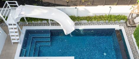 A swimming pool with a white slide, surrounded by a white wall and a wooden deck with chairs.