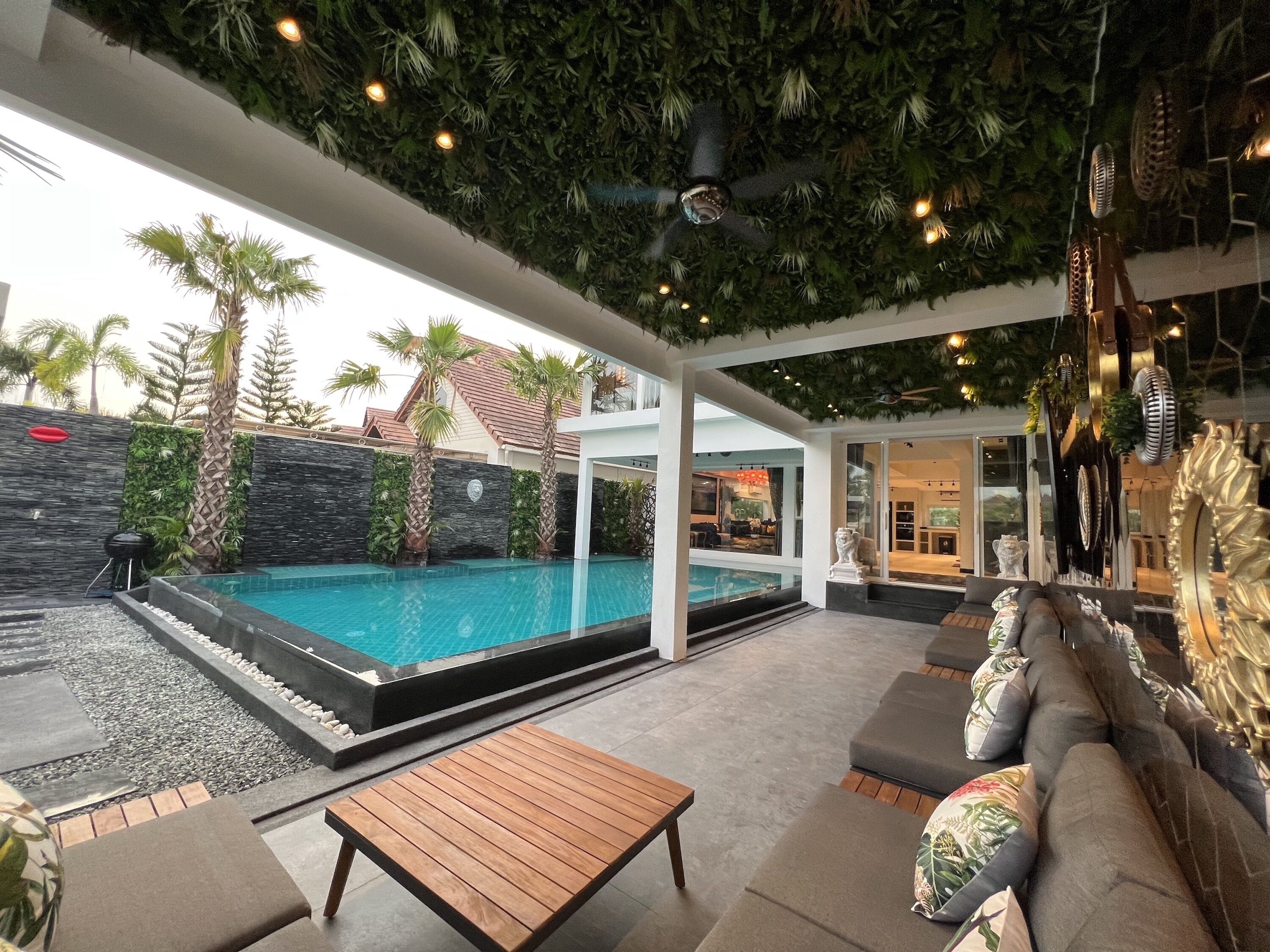 A modern outdoor pool area with a wooden table and seating, surrounded by palm trees and a stone wall.