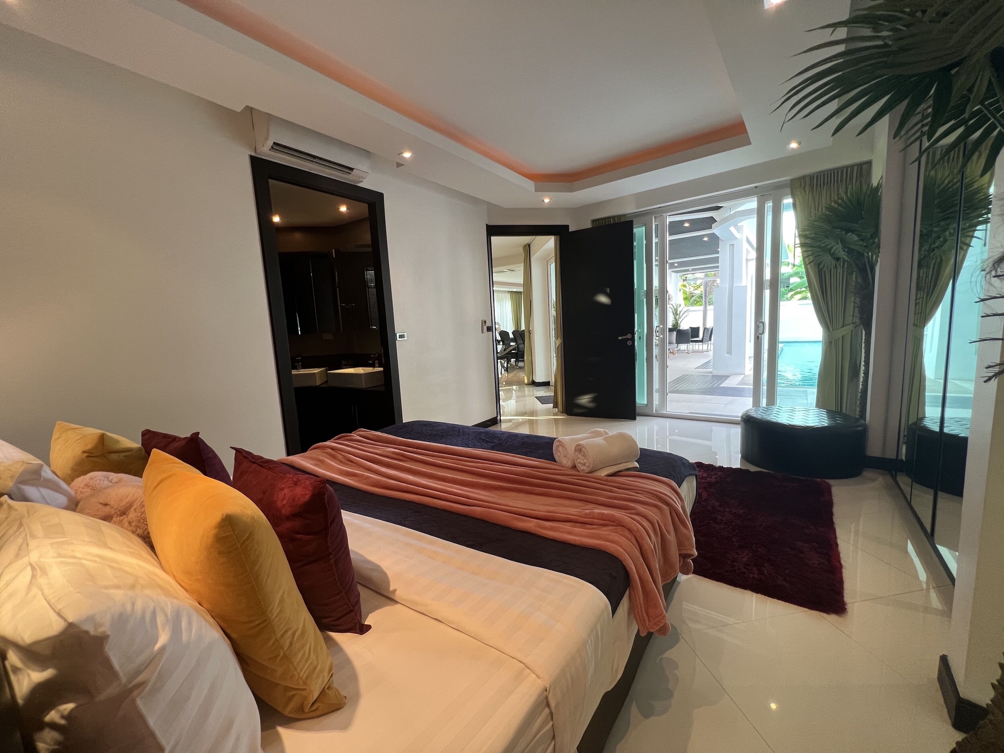 A modern hotel room with a large bed, multiple pillows, a built-in air conditioning unit, a sliding glass door leading to a balcony, and a view of the outdoors.