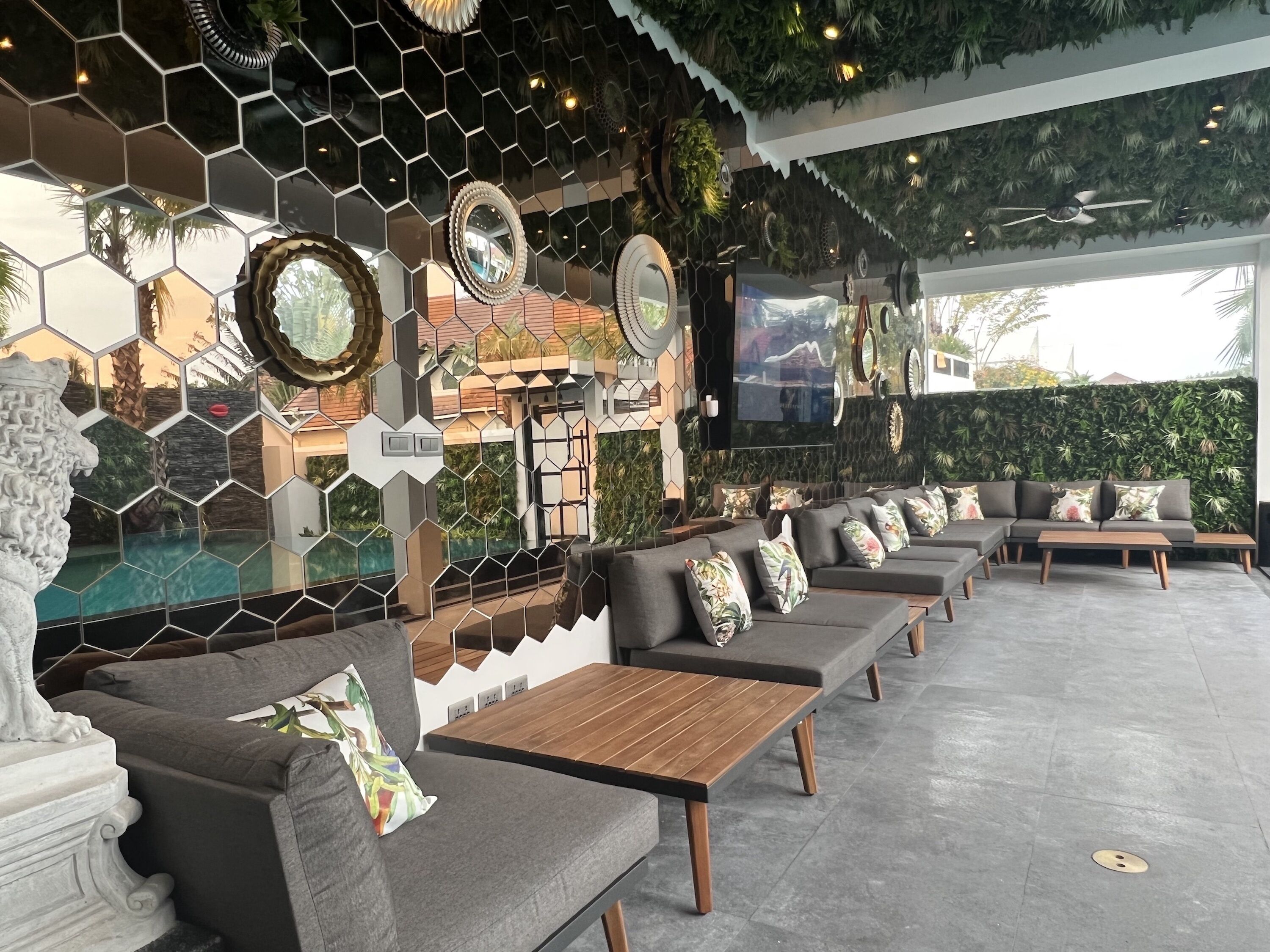 Uma área de estar moderna ao ar livre, com ladrilhos hexagonais, sofás acolchoados, mesas de madeira e espelhos decorativos.