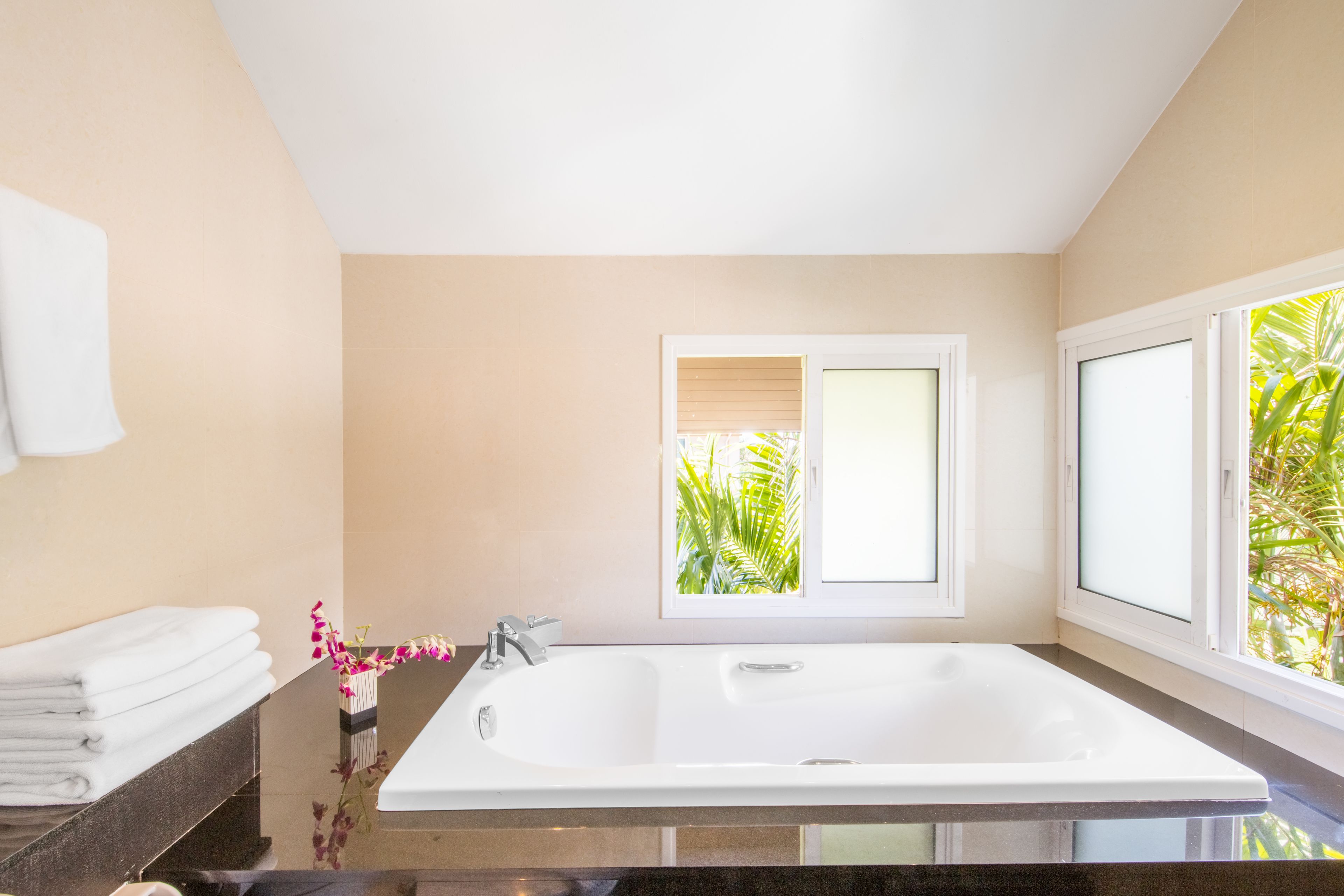 A modern bathroom with a large bathtub, a window with a view of greenery, and a stack of white towels on a counter.