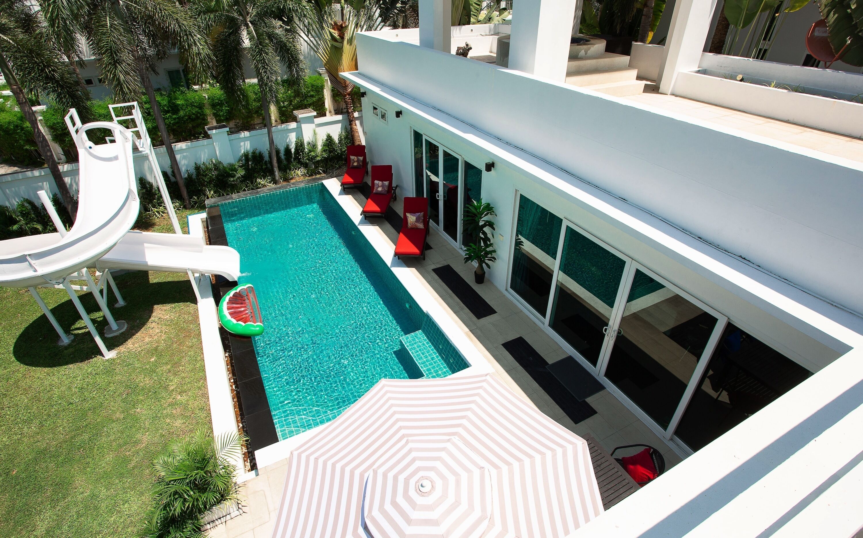 A modern outdoor swimming pool with a slide, red lounge chairs, and a large umbrella.