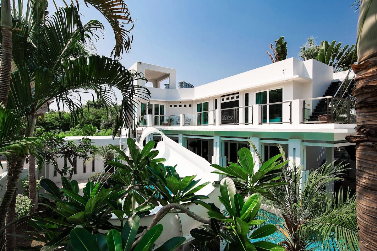 Une maison moderne à deux étages avec une piscine, entourée d’une végétation tropicale luxuriante.