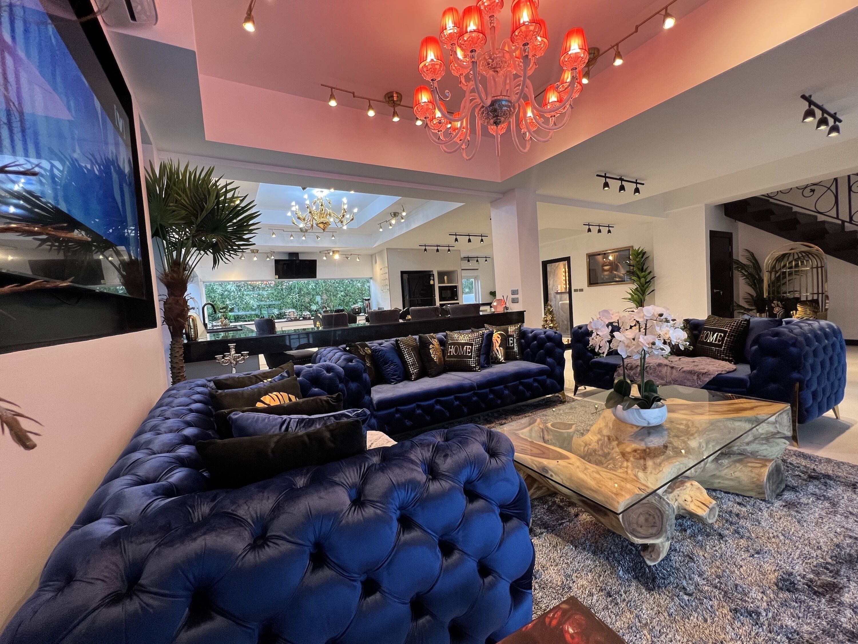 A spacious living area with a large sectional sofa, a glass coffee table, and a chandelier. There is a kitchen area visible in the background with a large window.