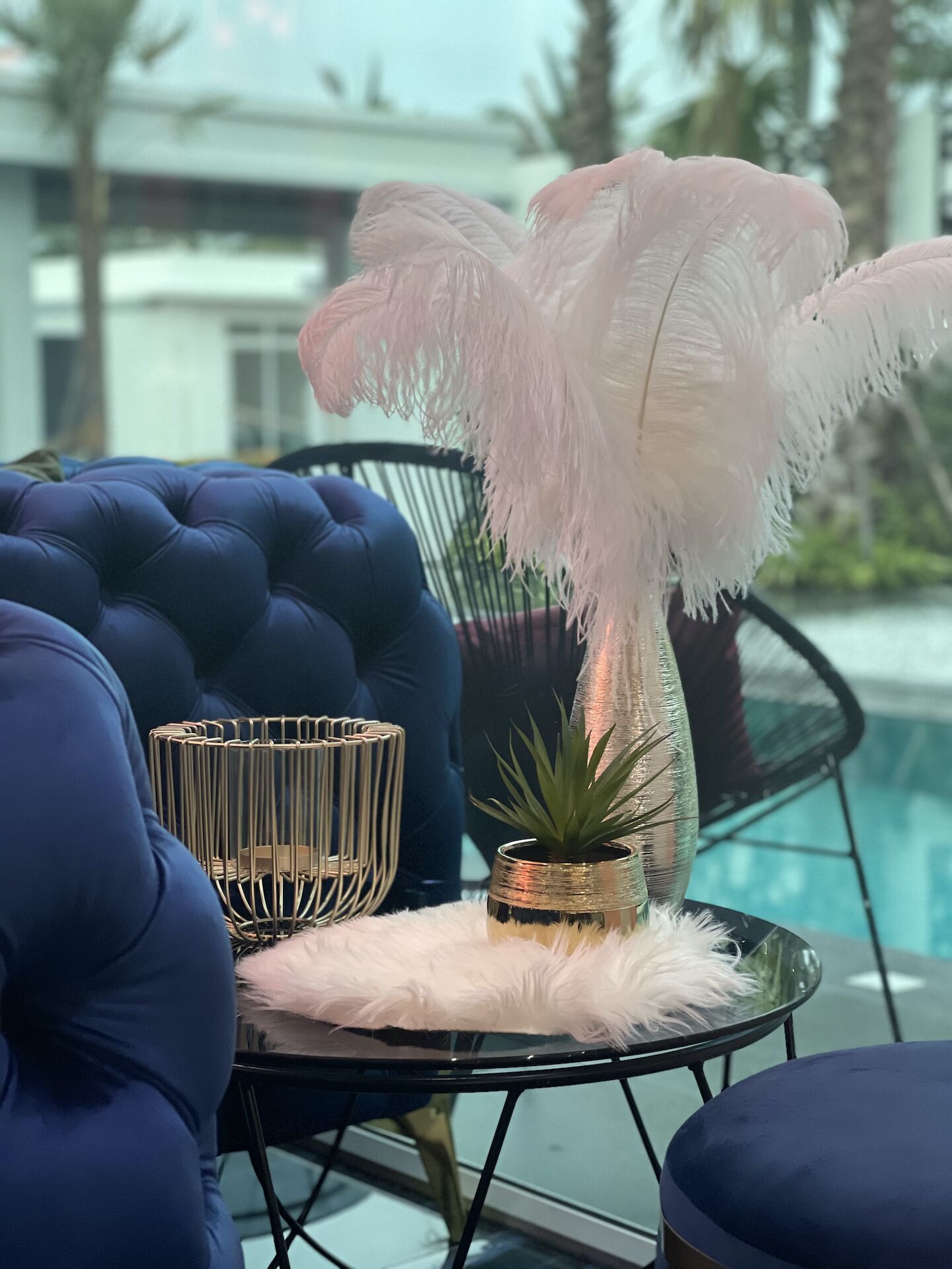 A modern outdoor seating area with a glass table, a fluffy white cushion, a small potted plant, and a decorative metal vase.