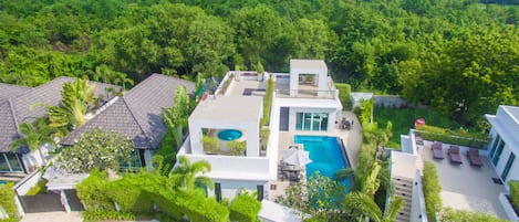 A modern house with a swimming pool, surrounded by lush greenery and a city skyline in the background.