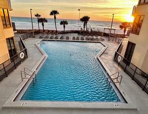 A rectangular pool with 8 ft. depth markers, surrounded by lounge chairs and palm trees, with a view of the ocean and sunset. - Sunbird 210w-Cozy Beachfront Condo with Sunset Views – Prime PCB Location (Panama City Beach)