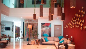 Een ruime lobby met een hoge plafond, een rode accentmuur, en een moderne zithoek met een bank en ronde houten tafels.