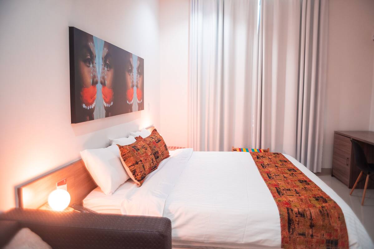 Een net opgemaakt bed met witte lakens, een bedektje met een patroon en een nachtkastje met een lamp.