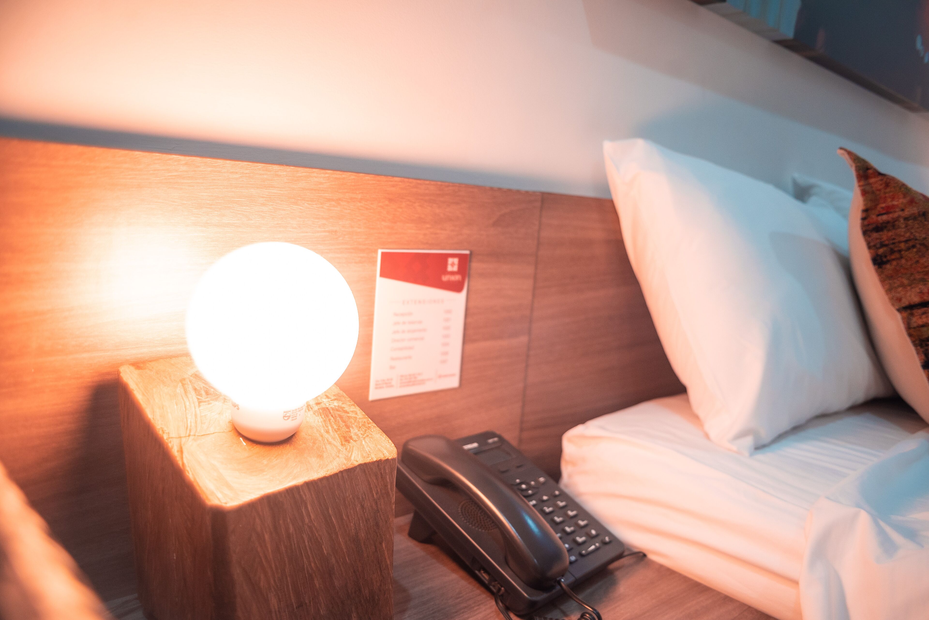 Una habitación de hotel con una lámpara de noche, un teléfono y una tarjeta de bienvenida en la pared.