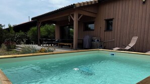 Ein rechteckiger Swimmingpool mit einer umlaufenden Holzterrasse, ein überdachter Außenbereich mit Tisch und Stühlen und im Hintergrund ein Holzhaus mit Fenstern und Schornstein.