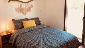 Ein Schlafzimmer mit einem gemusterten Bettüberwurf, einem gelben Kopfkissen, einer Wanddeko aus Makramee und einer Pendelleuchte.