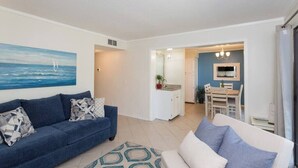 A living room with a blue sofa and a white sofa, a painting of the sea, and a kitchen area in the background. - 143 By the Sea * Gated Myrtle Beach Resort * 2/2 (Horry County)