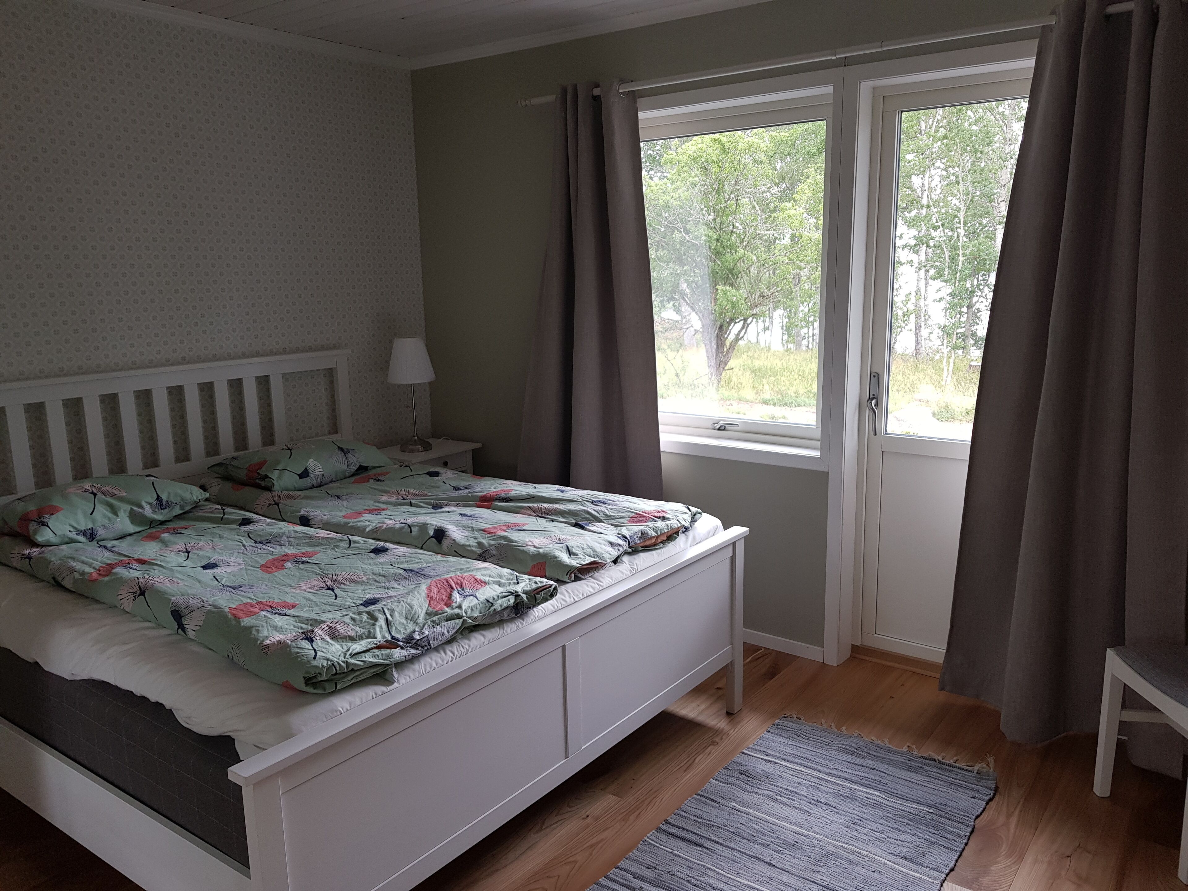 A bedroom with a bed, a nightstand, a window with curtains, and a view of trees outside.