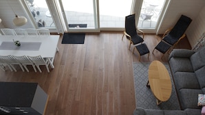 A modern living room with a wooden floor, a gray sofa, a dining table with chairs, and a large window offering a view of the outdoors. - Lövanäs Newly built modern house in a secluded location directly on the lake with its own jetty, boat (Korsberga)