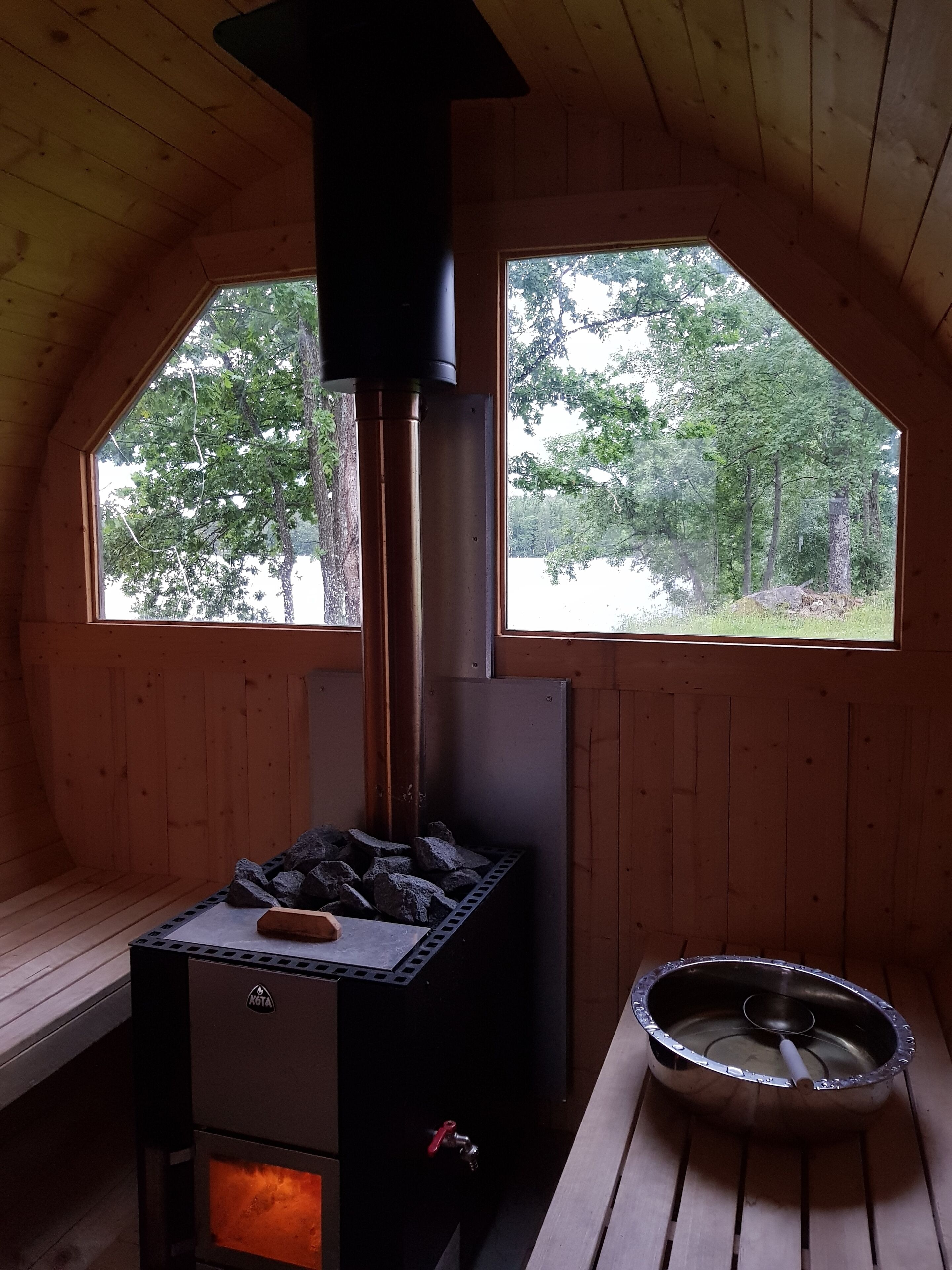 En mysig stuga med en vedeldad kamin, en metallskål och två fönster med utsikt över skogen.
