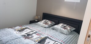 Un lit avec une couvre-lit à motifs et deux oreillers, une table de chevet avec une lampe et une prise électrique au mur.