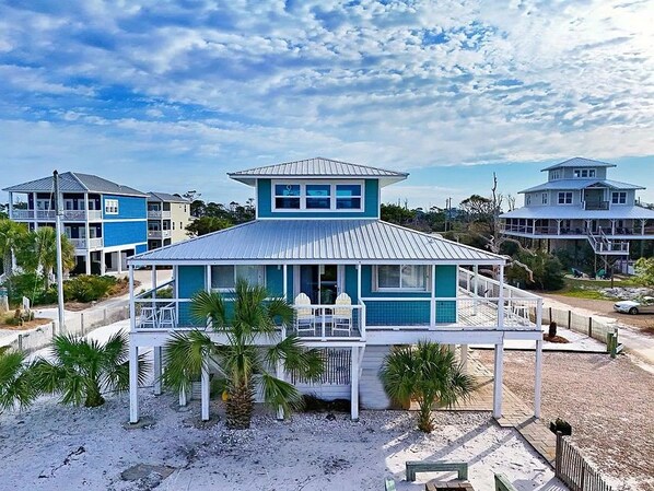 A beachfront house with a prominent tower, a covered porch, and a sandy beach area.