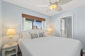 A bedroom with a bed, two bedside lamps, a ceiling fan, a window with blinds, and a door.