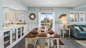 A dining area with a wooden table set for six, white chairs, and a view of the beach through a glass door.