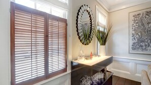 A hallway with wooden blinds, a mirror, a vase with plants, and a framed artwork on the wall. - 1828 Cottage | Heated Pool in Private Garden | Sleeps 6 | Pets | On-Site Parking (Savannah)