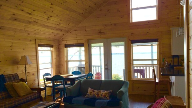 A wooden cabin interior with a sofa, a chair, a table, and a view of the water through large windows. - Charlottetown Hrbr’view Executive Waterfront (Stratford)