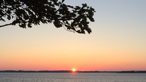 A sunset over a calm body of water with a grassy shoreline and a wooden structure in the foreground. - Charlottetown Hrbr’view Executive Waterfront (Stratford)