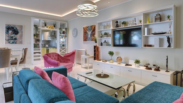 Una sala de estar moderna con un sofá, un sillón y un televisor. Se observan estanterías con objetos decorativos y una zona de comedor al fondo.
