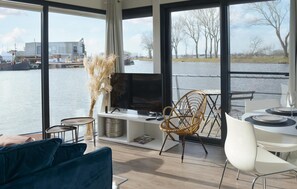 Ein modernes Wohnzimmer mit großem Fenster mit Flussblick, einem Flachbildfernseher, einer Essecke mit weißen Stühlen und einem blauen Sofa.