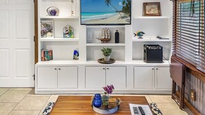 Ein Wohnzimmer mit an der Wand montiertem Fernseher, einem Couchtisch aus Holz mit Dekoration und einer weißen Schrankwand mit Regalen, die Bücher, ein Schiffmodell und eine Zimmerpflanze beherbergen.
