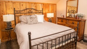 Un dormitorio con estructura de cama metálica, cabecera de madera, mesita de noche con lámpara, cómoda y espejo.