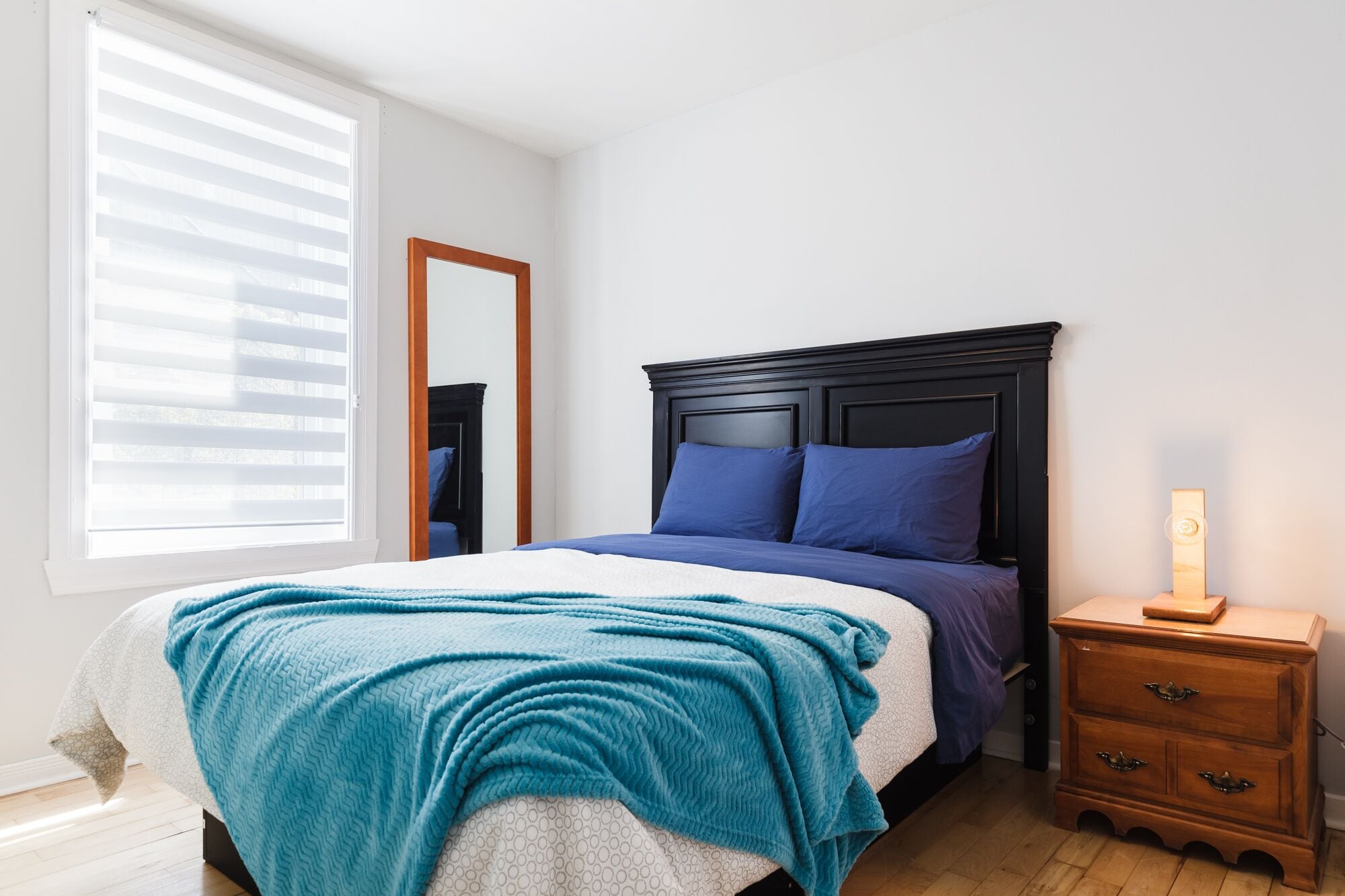 Ein Schlafzimmer mit einem Bett, einem Nachttisch, einem Spiegel und einem Fenster mit Jalousien.