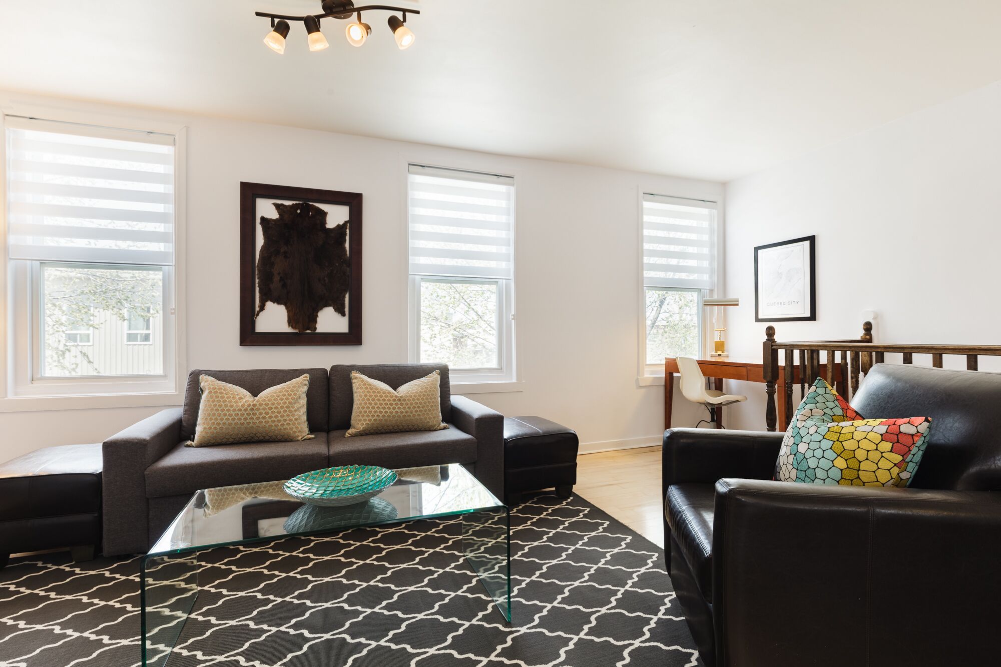 Sala de estar con sofá, sillón y una mesa de centro. Hay dos ventanas con persianas, un cuadro enmarcado en la pared y un escritorio con una silla.