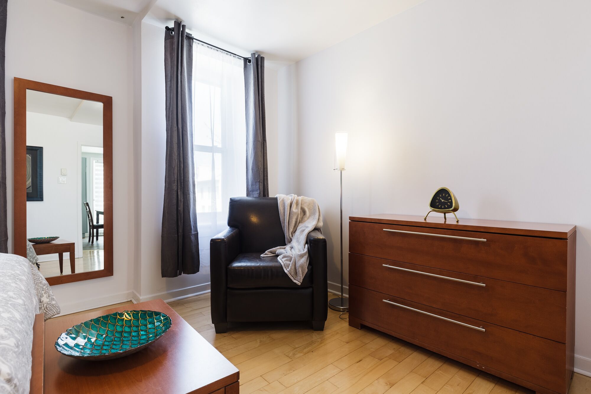 Une chambre à coucher avec un lit, une chaise, une commode, un miroir et une fenêtre avec des rideaux.