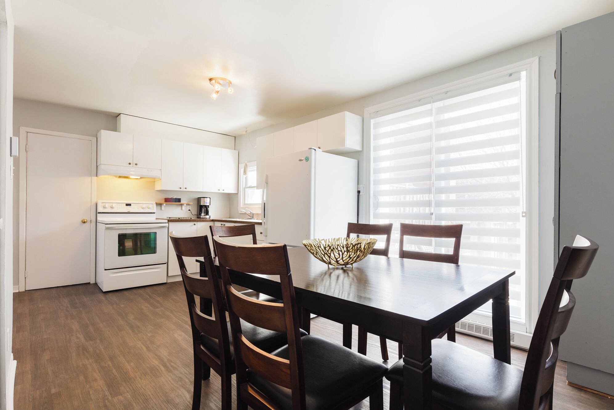 A compact kitchen with white cabinets, a stove, and a coffee maker. There is a dining table with four chairs and a decorative bowl.