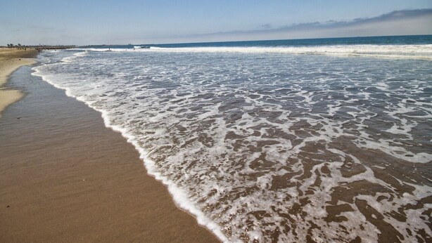 A beach with waves gently lapping onto the shore, a clear sky, and a distant shoreline with people and structures. - Charming Ventura Beach Bungalow on Prime Beach Lane in Pierpont by the Sea (Ventura)