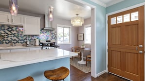 A kitchen with white cabinets, a marble island, wooden stools, a chandelier, and a wooden door. - Charming Ventura Beach Bungalow on Prime Beach Lane in Pierpont by the Sea (Ventura)