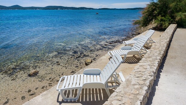 Une plage à l’eau claire, avec des chaises longues blanches et un mur de pierre.