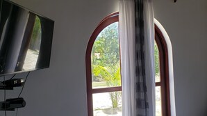 Ein Zimmer mit an der Wand befestigtem Fernseher, einem Fenster mit Blick auf Grünfläche und einem teilweise zurückgezogenen Vorhang.